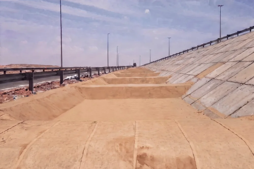 A drainage channel graded into bare sand beside a highway, with an older concrete slab revetment on the bank above it