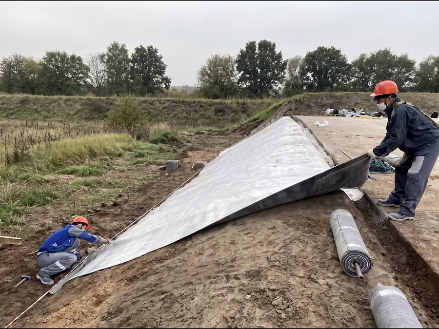 Two workers pulling a sheet of the material out over a prepared sand slope, a roll lying on the sand beside them