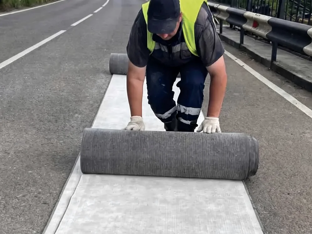 A worker pushing out a roll along a road, dark dry material behind and pale cured surface ahead